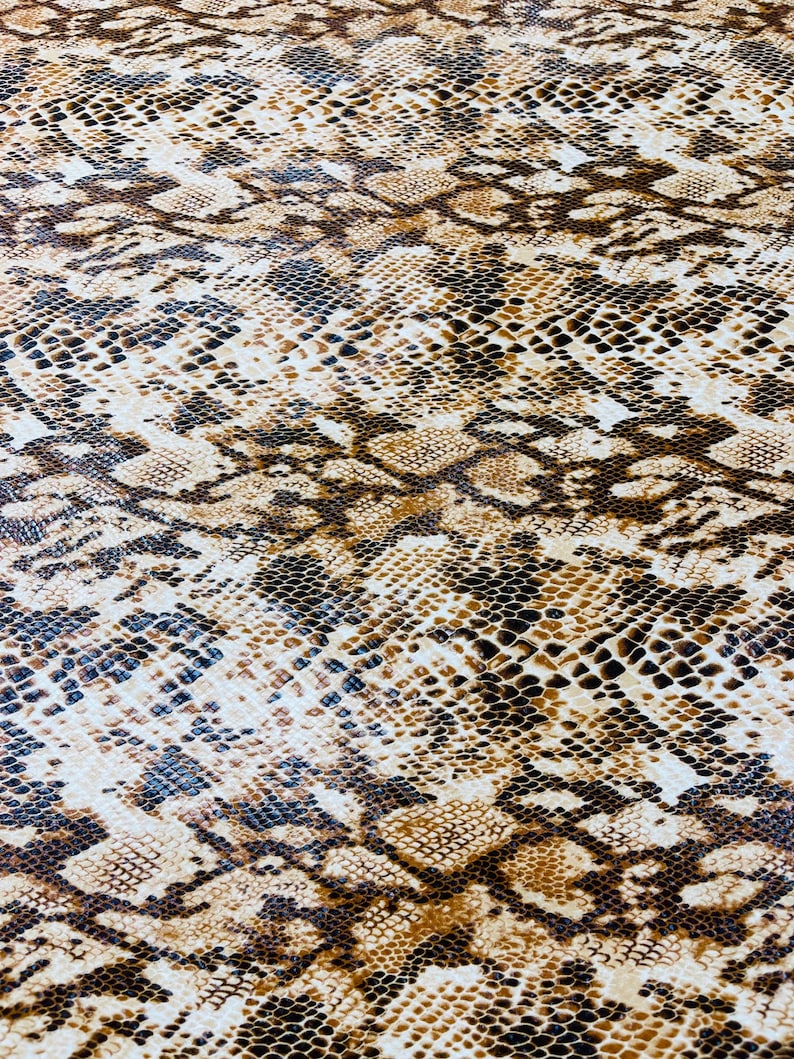 Pu&ograve; includere: Un primo piano di un tessuto con motivo a pelle di serpente marrone e bianco. Il tessuto ha una superficie testurizzata e un motivo a scaglie ripetuto.