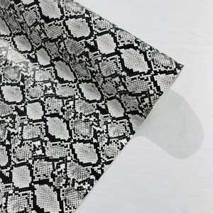 May include: A roll of material featuring a high-contrast black and white snakeskin pattern. The surface displays a repeating, textured scale design with varying shapes and sizes of black outlines against a white background, shown partially unrolled.