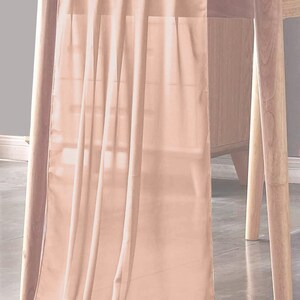 May include: A peach-colored table runner draped over a wooden table. The fabric is sheer and flowing, with vertical folds creating a textured effect. The table has light-colored wooden legs and a matching tabletop.
