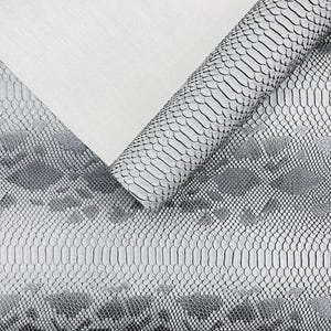 May include: A roll of faux leather with a snakeskin pattern in shades of gray and white. The material is partially rolled, revealing the underside, which is a plain white. The snakeskin design features a scale-like texture.