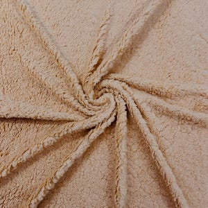 May include: Close-up of a beige sherpa fabric with a soft, textured surface. The fabric is arranged in a spiral pattern, with visible seams and a plush, fuzzy texture. The color is a warm, neutral tone.