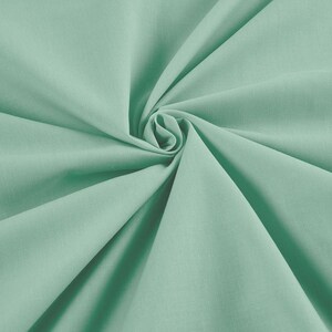 May include: A close-up shot of a swirl of mint green fabric. The fabric is smooth and appears to be a lightweight material, possibly linen or cotton. The folds create shadows and highlights, adding depth to the image. The color is a soft, muted green.