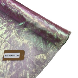 May include: A roll of iridescent metallic tissue lame fabric with a green, purple, and gold color scheme. The fabric has a crinkled texture and a label that reads "Metallic Tissue Lame".