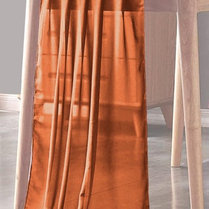May include: A burnt orange table runner made of a soft, flowing fabric. The runner is draped over a wooden table and hangs down to the floor.
