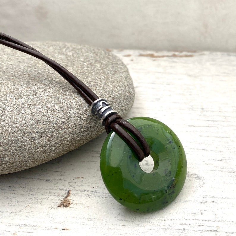 high quality Canadian nephrite dark green jade pendant, a 30mm donut shaped gemstone, threaded on a black leather cord, with a handmade sterling silver zodiac libra symbol bead sitting above the stone,  against a grey pebble, white wood background.