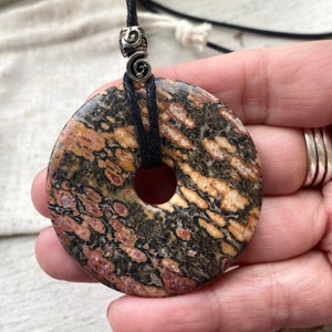 Puede incluir: Un colgante redondo de piedra negra, marrón y roja con un agujero en el centro. El colgante está en un cordón negro con cuentas de metal plateado.