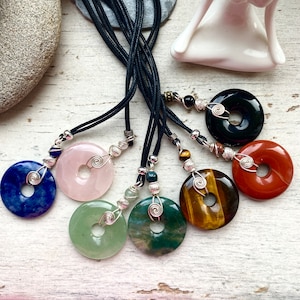 Può includere: Sette collane con ciondoli in pietra di diversi colori. Ogni ciondolo è un disco rotondo con un foro al centro. Le pietre sono blu, rosa, verdi, marroni, rosse, nere e verde screziato. Le collane sono realizzate con cordoncino nero e filo d'argento.