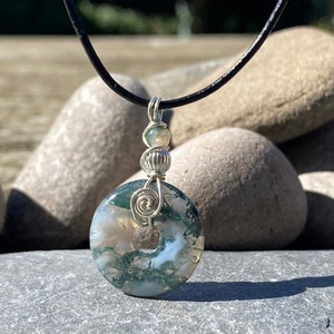 May include: A pendant necklace with a moss agate stone, displaying green and white patterns, wire-wrapped in silver. The stone is round with a central hole and hangs from a black leather cord. A unique piece of jewellery.