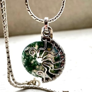 Puede incluir: Un collar con colgante de plata con una piedra redonda de ágata musgosa, que muestra patrones verdes y blancos. La piedra está engastada en un marco de alambre de plata con un diseño decorativo. La cadena del collar también es plateada.