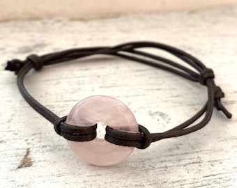 May include: A pink rose quartz gemstone charm bracelet with a brown leather cord. The cord is tied with a knot on each end.