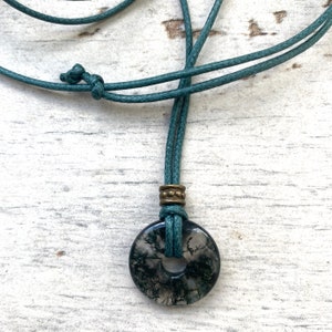 May include: A dark green cord necklace with a round, black and white agate pendant. The pendant has a hole in the centre and is accented with a brass bead.