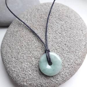 Op de afbeelding: Een groene jade hanger in de vorm van een cirkel met een gat in het midden, hangend aan een zwarte koordsnoer. De hanger ligt op een grijze steen.