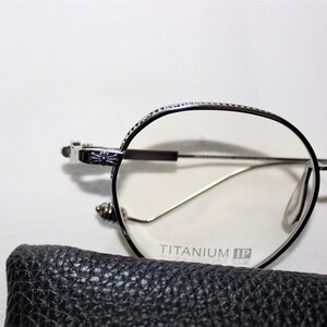 May include: Silver-toned round eyeglasses with a cross detail on the side. The glasses have a clear lens and the words "TITANIUM IP" are printed on the frame.