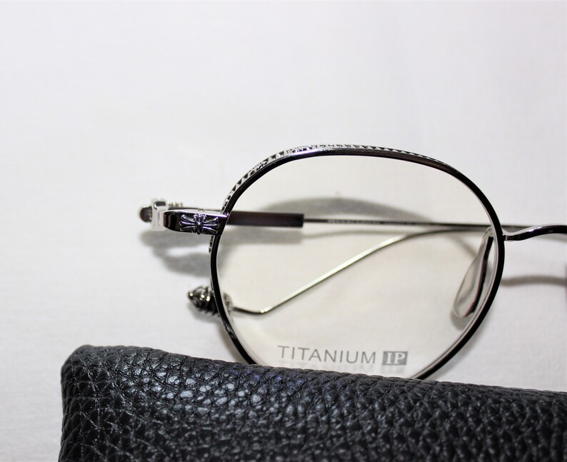May include: Silver-toned round eyeglasses with a cross detail on the side. The glasses have a clear lens and the words "TITANIUM IP" are printed on the frame.