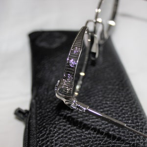 May include: Silver-toned metal eyeglasses with a decorative design on the arms. The glasses are resting on a black leather case.