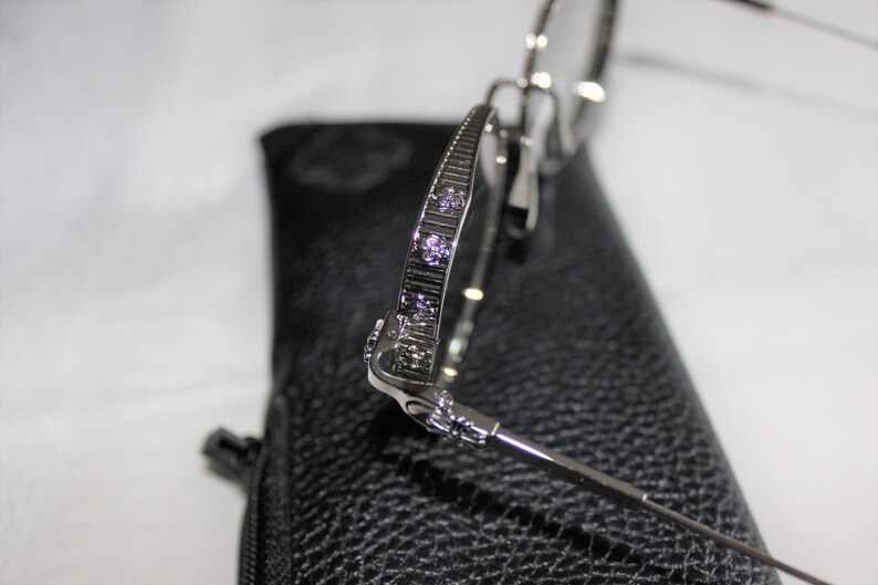 May include: Silver-toned metal eyeglasses with a decorative design on the arms. The glasses are resting on a black leather case.