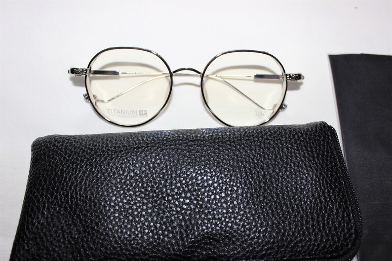 May include: Silver-rimmed round eyeglasses with clear lenses and a black leather glasses case. The glasses have the text "Titanium" printed on the frame.