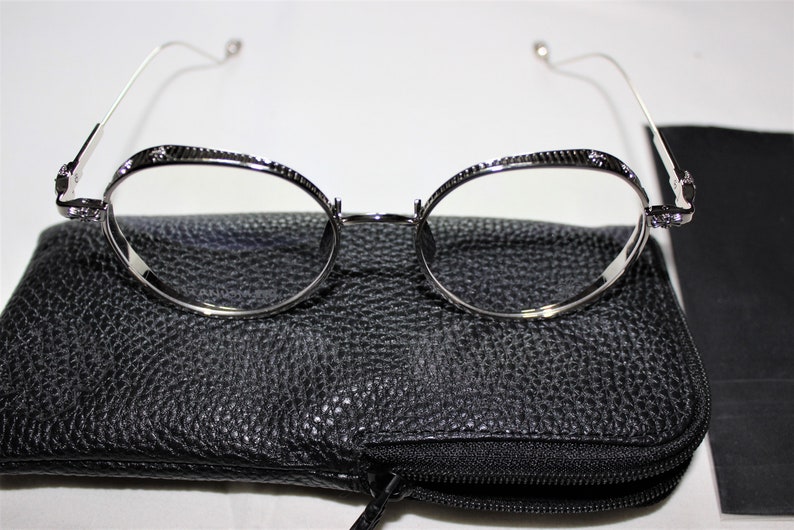 May include: Silver round-framed glasses with a black leather case. The glasses have a textured metal frame and clear lenses.