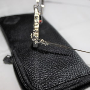 May include: Silver sunglasses with a detailed cross design on the arms, resting on a black leather case with a zipper.