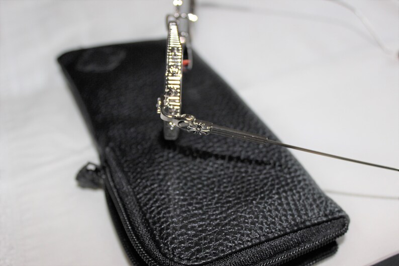 May include: Silver sunglasses with a detailed cross design on the arms, resting on a black leather case with a zipper.