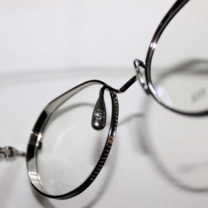 May include: Silver-toned metal eyeglasses with round frames and a thin metal nose bridge. The glasses have a decorative pattern on the arms.