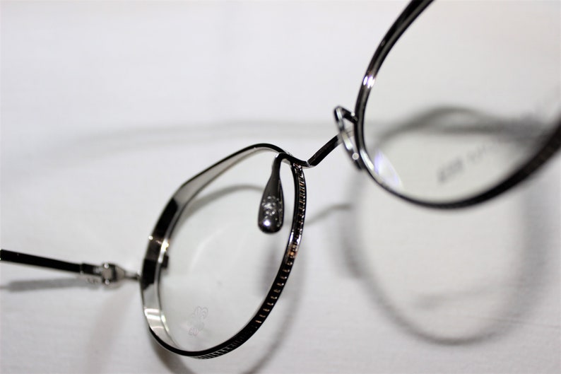 May include: Silver-toned metal eyeglasses with round frames and a thin metal nose bridge. The glasses have a decorative pattern on the arms.