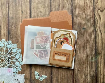 Op de afbeelding: Een verzameling vintage ephemera, waaronder postzegels, labels en een "Admit One"-ticket, gerangschikt in een bruine map. Decoratieve elementen zijn een lichtblauw kantkleedje en een vlindersticker, alles tegen een houten achtergrond.