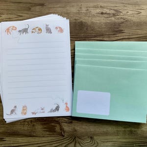 Può includere: Una pila di fogli di carta da lettere a righe con illustrazioni di gatti ad acquerello in arancione, grigio e marrone. Accanto, una pila di buste verde chiaro con un'area etichetta bianca. La cancelleria è su una superficie di legno.
