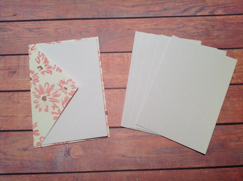 Puede incluir: Una tarjeta blanca con un sobre de patr&oacute;n floral. El sobre est&aacute; doblado sobre la tarjeta. Hay cinco tarjetas blancas en blanco apiladas en el lado derecho de la imagen.