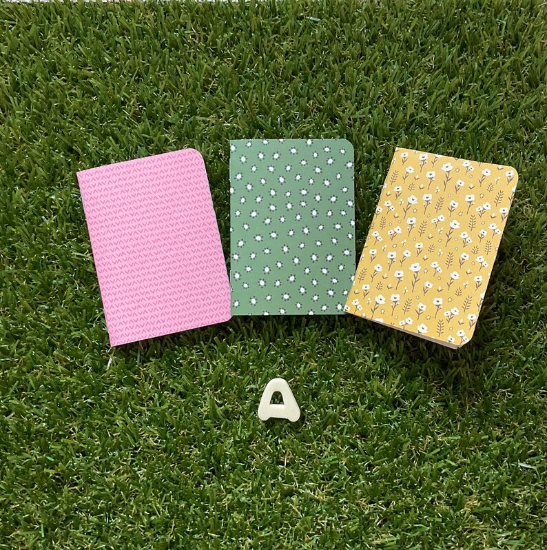 May include: Three small notebooks with different patterned covers. The notebooks are pink with a small white pattern, green with a small white flower pattern, and yellow with a small white flower pattern. The notebooks are arranged in a triangle shape on a green grassy surface.