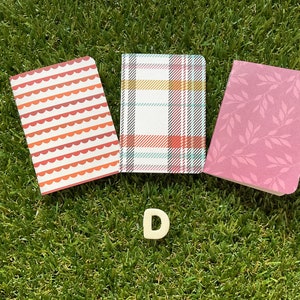 May include: Three small notebooks with different patterned covers. The covers are red and white with a wavy pattern, white and black plaid, and pink with a leaf pattern. The notebooks are on a green grassy surface with a white letter D in the foreground.