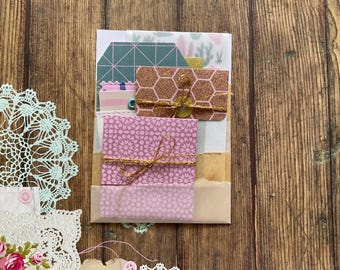 Op de afbeelding: Een verzameling decoratieve papieren ephemera, waaronder papier met patronen, kleine enveloppen en labels, vastgebonden met gouden touw. Het kleurenpalet omvat roze, goud en blauwgroen, met een houten achtergrond. De items zijn gerangschikt in een gelaagde compositie.