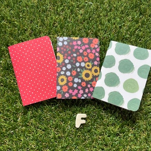 May include: Three small notebooks with different patterned covers. The first notebook has a red cover with white polka dots. The second notebook has a black cover with a floral pattern in shades of pink, yellow, and green. The third notebook has a white cover with green polka dots.