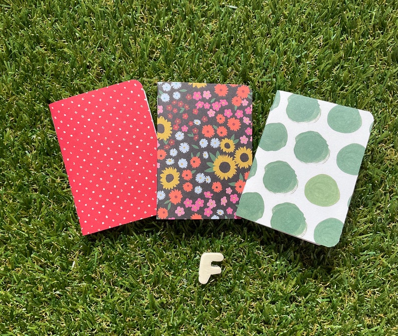 May include: Three small notebooks with different patterned covers. The first notebook has a red cover with white polka dots. The second notebook has a black cover with a floral pattern in shades of pink, yellow, and green. The third notebook has a white cover with green polka dots.