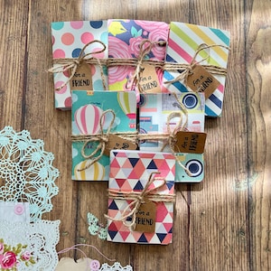 May include: A stack of eight small, colourful notebooks tied with twine and a brown paper tag that says "For a Friend". The notebooks have different patterns, including polka dots, flowers, stripes, hot air balloons, and geometric shapes.