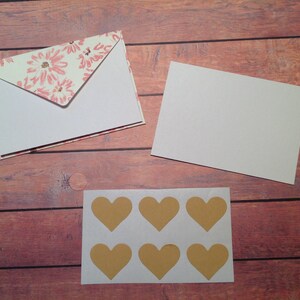 Puede incluir: Un conjunto de seis pegatinas doradas en forma de coraz&oacute;n sobre una hoja de papel blanca. Las pegatinas est&aacute;n dispuestas en dos filas de tres. Las pegatinas est&aacute;n sobre un fondo de madera.