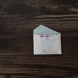May include: A small, white envelope with a colorful cartoon design on the front. The envelope is lying flat on a dark brown wooden surface.