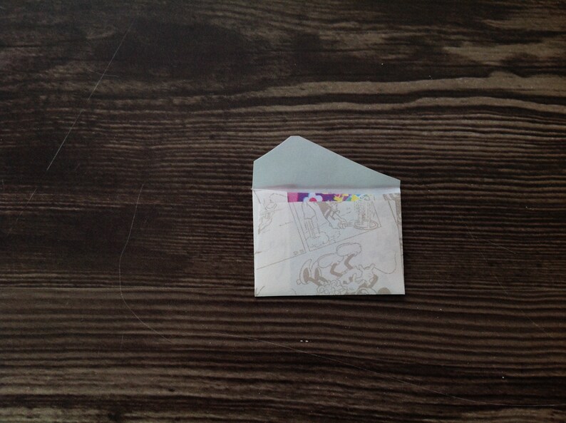 May include: A small, white envelope with a colorful cartoon design on the front. The envelope is lying flat on a dark brown wooden surface.