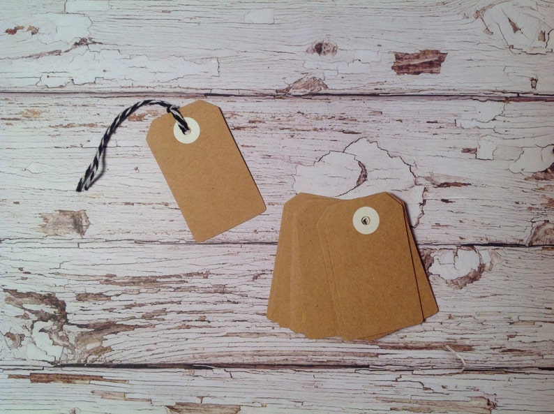 May include: A set of brown paper gift tags with white eyelets and twine. The tags are on a whitewashed wooden surface.