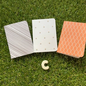 May include: Three small notebooks with different patterned covers. The covers are striped, dotted, and have a scallop design. The notebooks are on a green grassy surface.