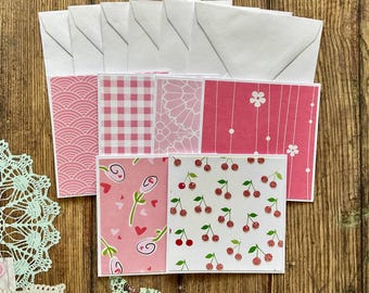 Pode incluir: Um conjunto de oito cartões de notas com padrões rosa e branco com envelopes correspondentes. Os cartões apresentam uma variedade de designs, incluindo xadrez, corações, cerejas e flores.