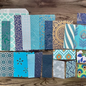 Op de afbeelding: Een verzameling papieren kaarten en enveloppen met patronen in tinten blauw, teal en goud. Ontwerpen omvatten bloemen-, geometrische en abstracte patronen. De kaarten zijn gerangschikt op een houten oppervlak en tonen een verscheidenheid aan texturen en stijlen.