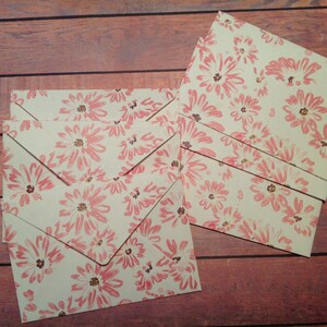 Puede incluir: Un conjunto de cinco sobres de color crema con un patr&oacute;n floral rosa. Los sobres est&aacute;n dispuestos en forma de abanico sobre una superficie de madera.