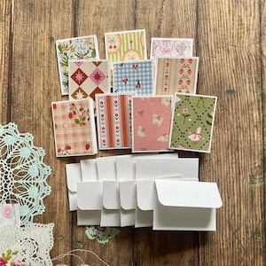 Puede incluir: Un conjunto de pequeñas tarjetas estampadas con sobres blancos a juego. Las tarjetas presentan varios diseños, incluyendo patrones florales, de cuadros y de fresas. La paleta de colores incluye rosa, verde, rojo y blanco, creando una estética vintage.