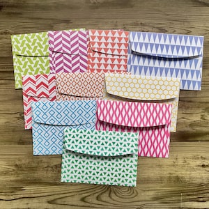 Pode incluir: Uma coleção de envelopes coloridos estampados com vários desenhos, incluindo ziguezagues, triângulos e formas geométricas. Os envelopes são em tons de verde, rosa, azul, laranja e amarelo, dispostos sobre uma superfície de madeira.