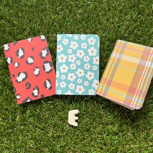 May include: Three small notebooks with different patterned covers. The first notebook has a red cover with black leopard print. The second notebook has a blue cover with white flowers. The third notebook has a yellow cover with a plaid pattern.