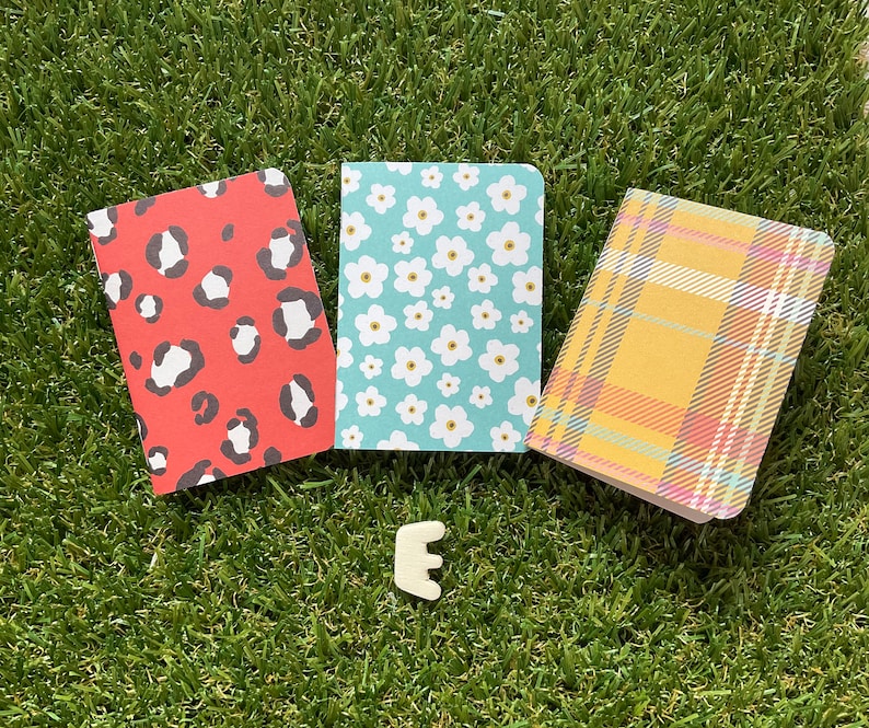 May include: Three small notebooks with different patterned covers. The first notebook has a red cover with black leopard print. The second notebook has a blue cover with white flowers. The third notebook has a yellow cover with a plaid pattern.
