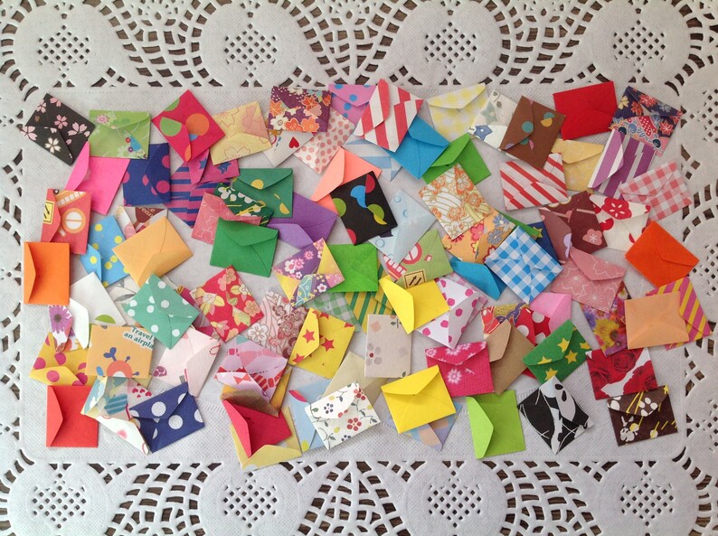 May include: A collection of colorful, patterned paper envelopes. The envelopes are small and are arranged in a random pattern on a white lace tablecloth.