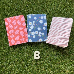 May include: Three small notebooks with different patterned covers. The covers are red with white leaf designs, blue with white flower designs, and red and white stripes.