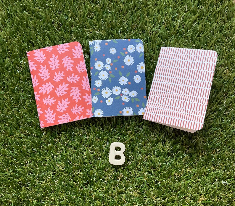 May include: Three small notebooks with different patterned covers. The covers are red with white leaf designs, blue with white flower designs, and red and white stripes.